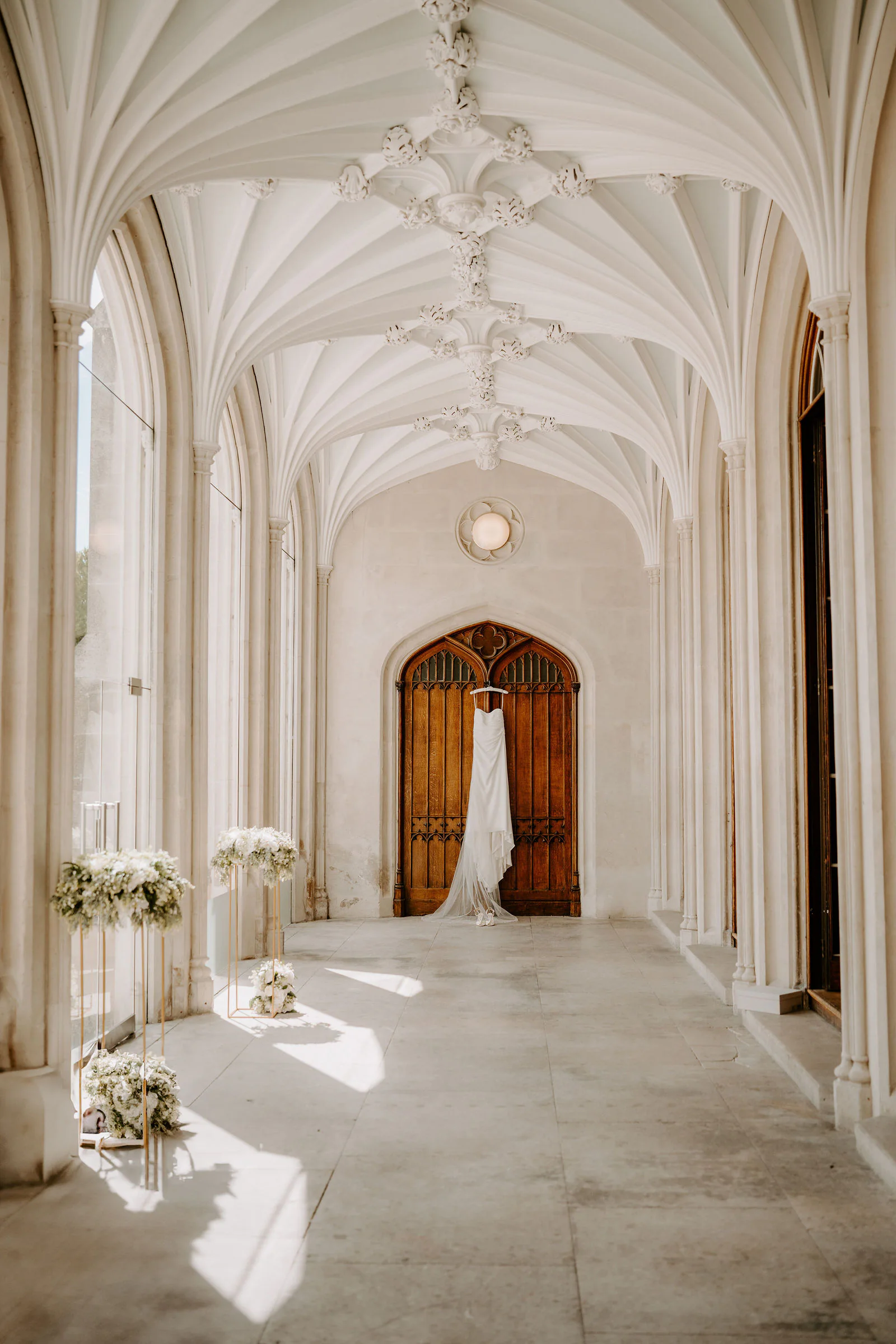 A wedding dress hangs from an arched wooden door at the end of a vaulted white stone cloister dressed with white flowers, with sunlight on the floor