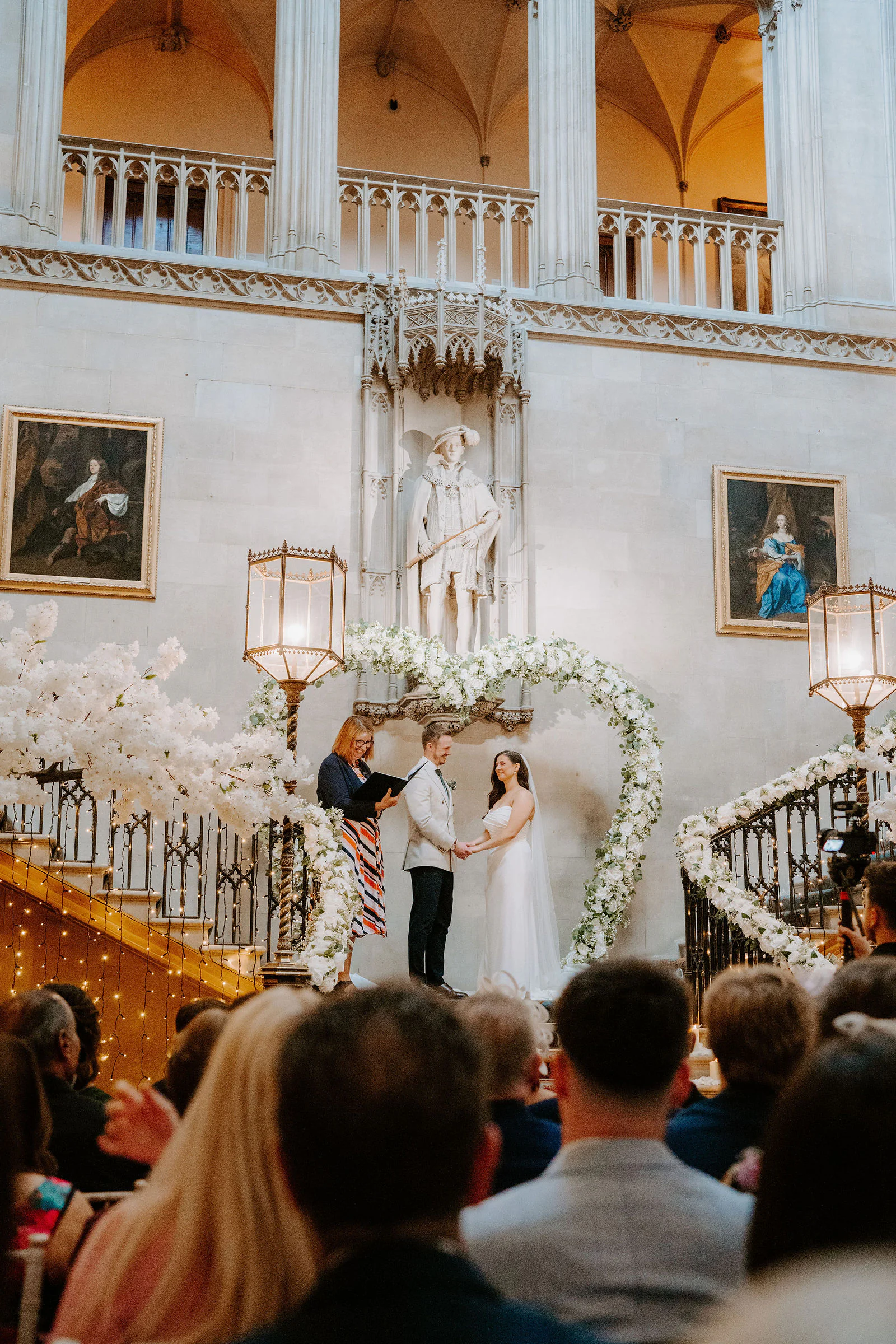 Phil and Natasha stand beneath a heart-shaped arch of white flowers at the foot of the grand staircase at Ashridge House as guests watch from the foreground