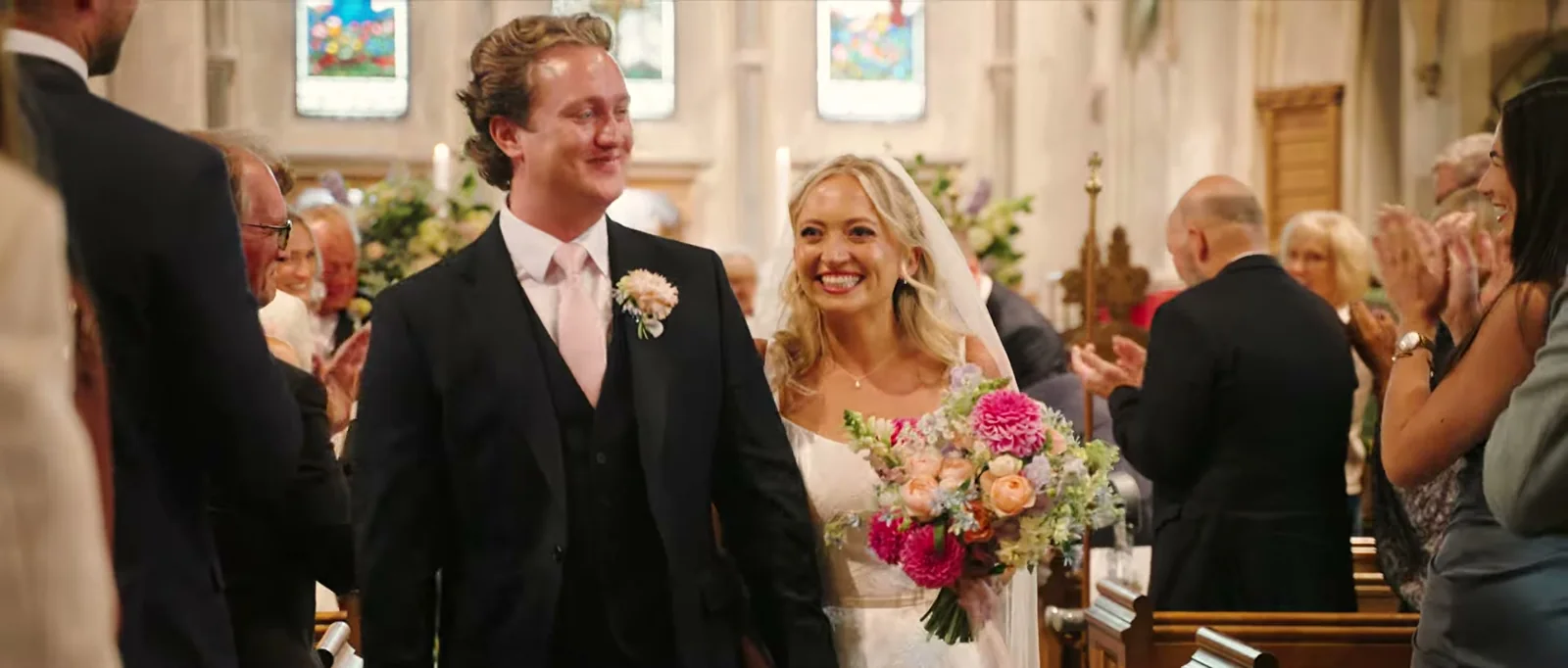 A groom and a bride with a bright bouquet walk up the aisle of a church as guests applaud