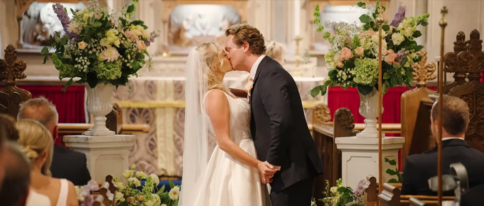 A couple kiss at the altar of a church beside tall arrangements of flowers