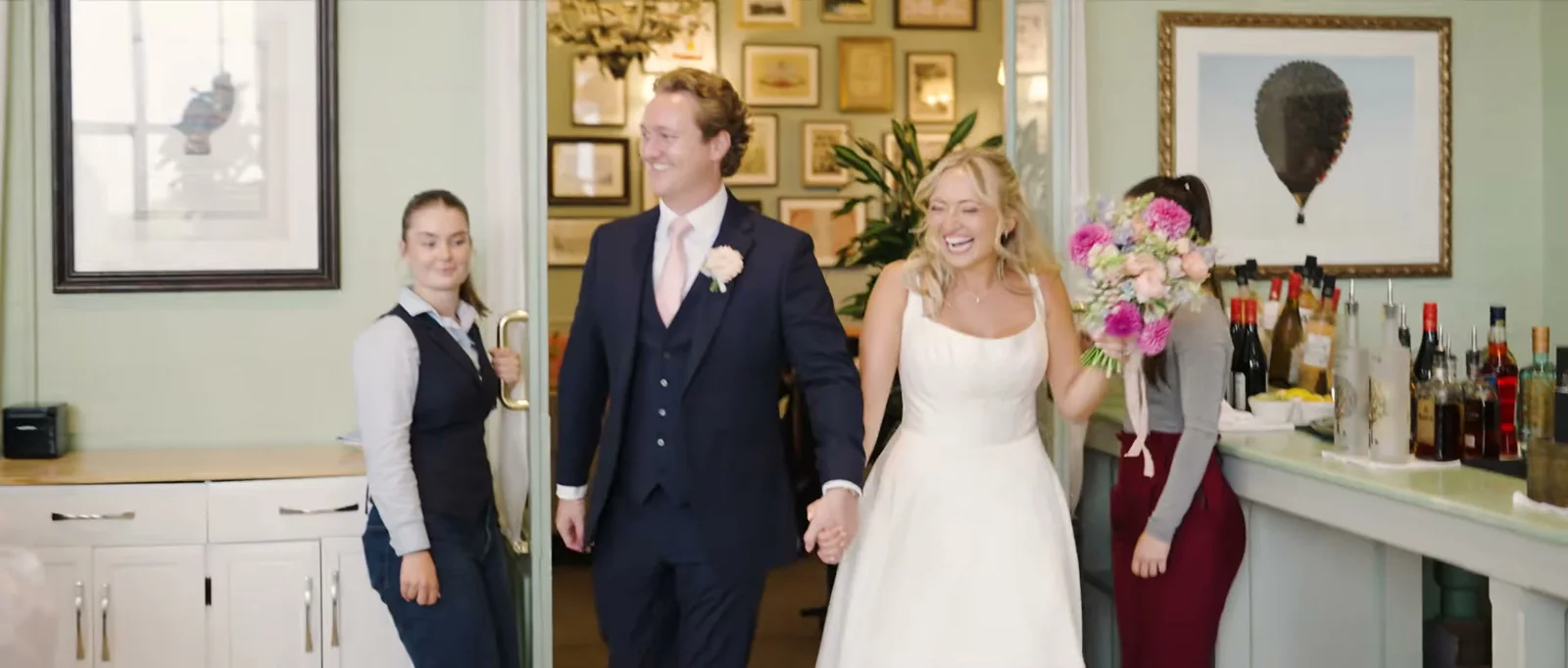 A couple hold hands as they walk past a bar where two members of staff smile at them