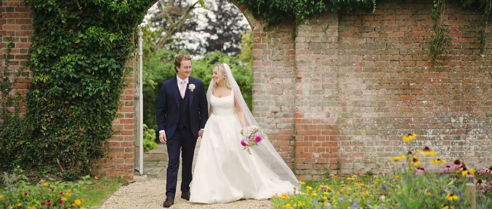 A bride and groom walk hand in hand through an arch in an old brick garden wall with wild flowers in the border
