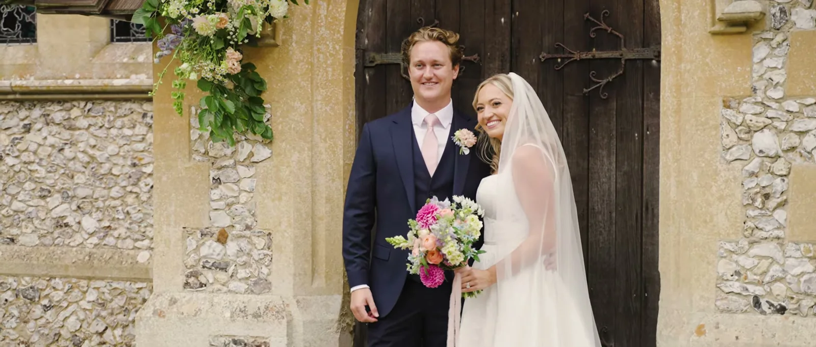 A couple stand smiling in front of the dark wooden door of a flint-walled church, the bride holding a bright bouquet