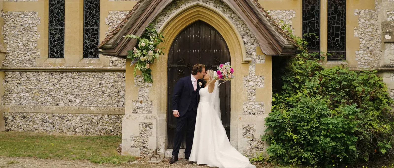 A couple kiss in the arched doorway of a small flint church framed by flowers