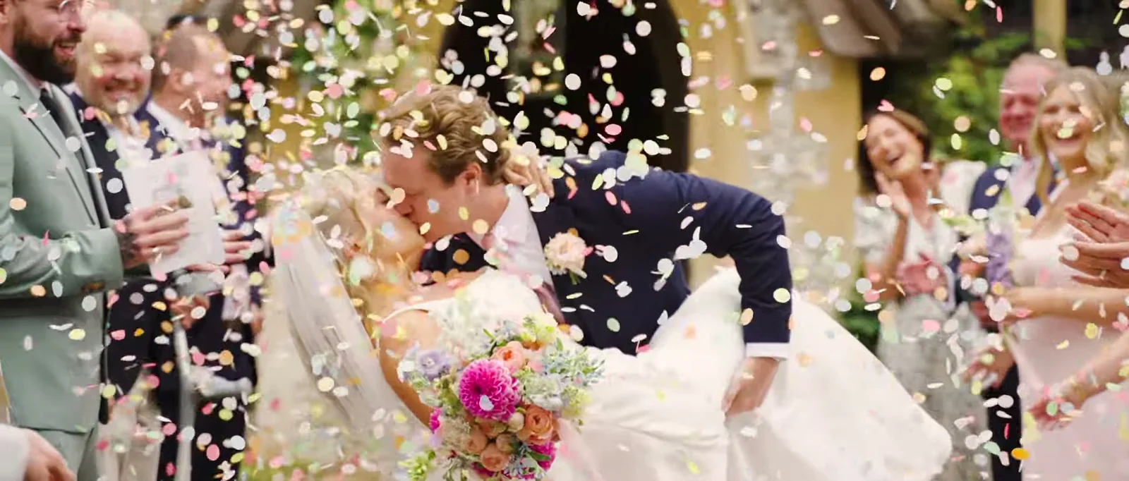 A couple kiss in a shower of confetti as guests laugh around them