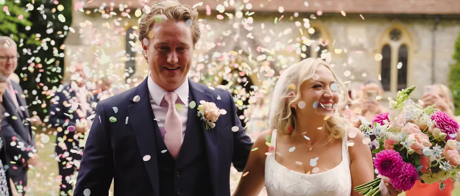 A groom and a bride smile as confetti falls around them outside a flint church