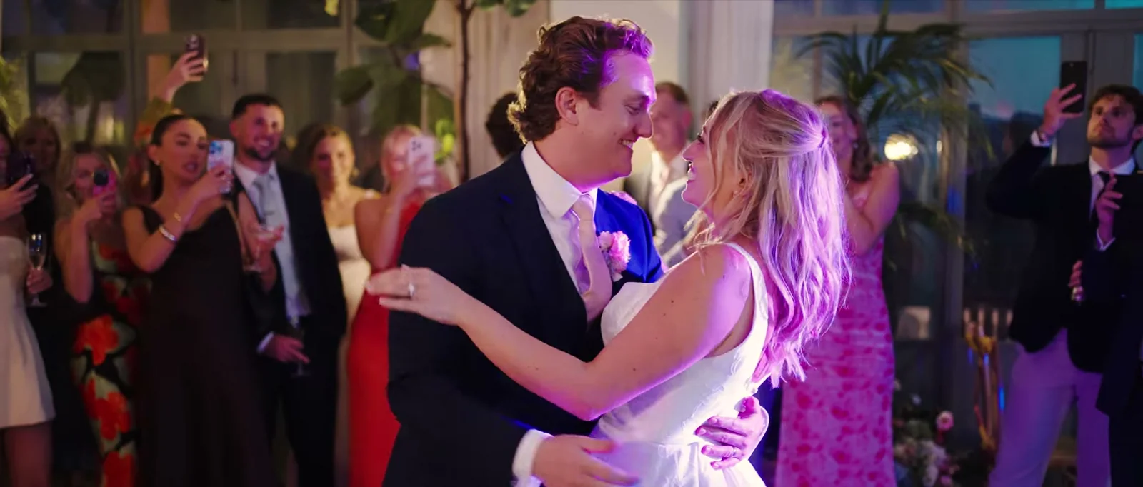 A groom and a bride share their first dance in a plant-filled room with guests watching