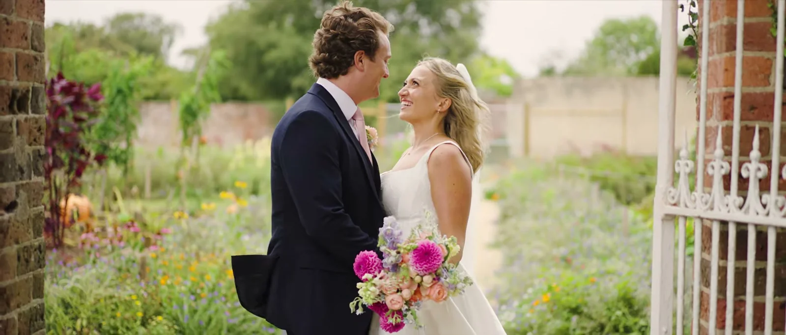 A groom in a navy suit and a bride in a veil laugh together beside a white iron gate in a walled garden full of flowers at The Retreat at Elcot Park