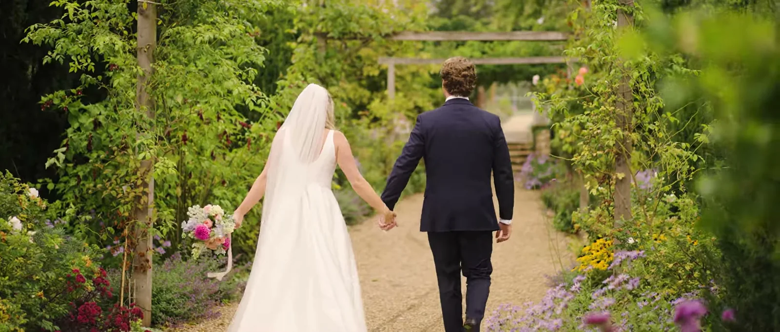 A bride in a long veil and a groom in a navy suit walk away hand in hand along a gravel path through a flower garden