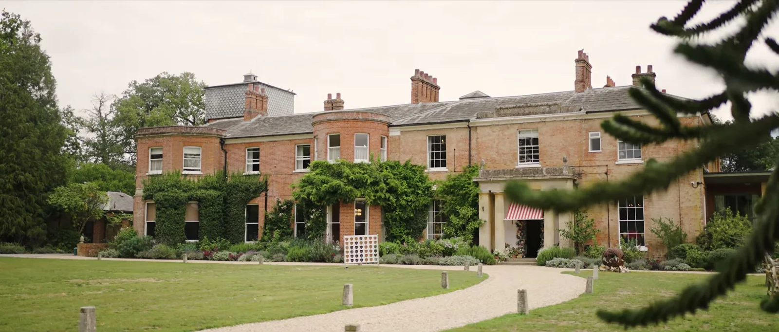The red-brick front of The Retreat at Elcot Park seen across a lawn, with climbing plants and a striped awning