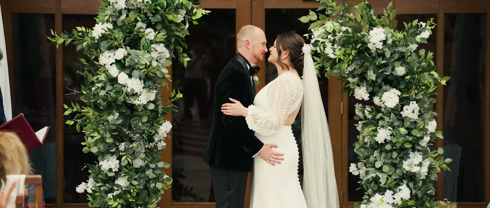 A groom and a bride in a veil are about to kiss in front of a tall arch of white flowers and greenery