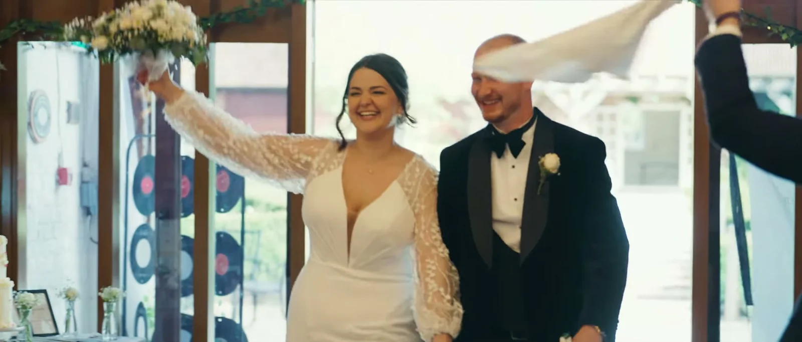 A bride raises her bouquet and a groom laughs as a guest waves a napkin over their heads in a doorway