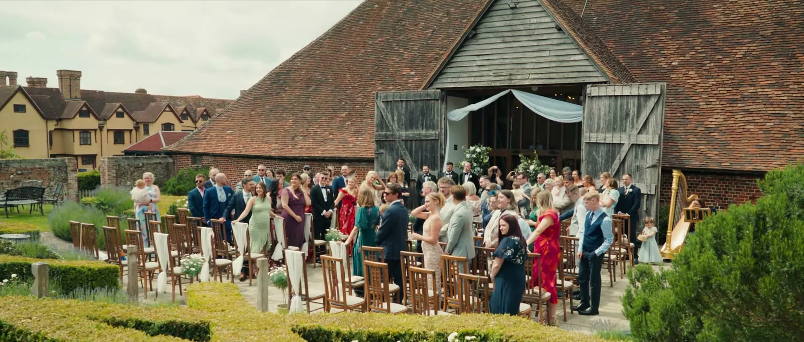 Guests stand beside rows of wooden chairs in a clipped box garden in front of the open doors of a big red-tiled barn, with a harp beside the doors