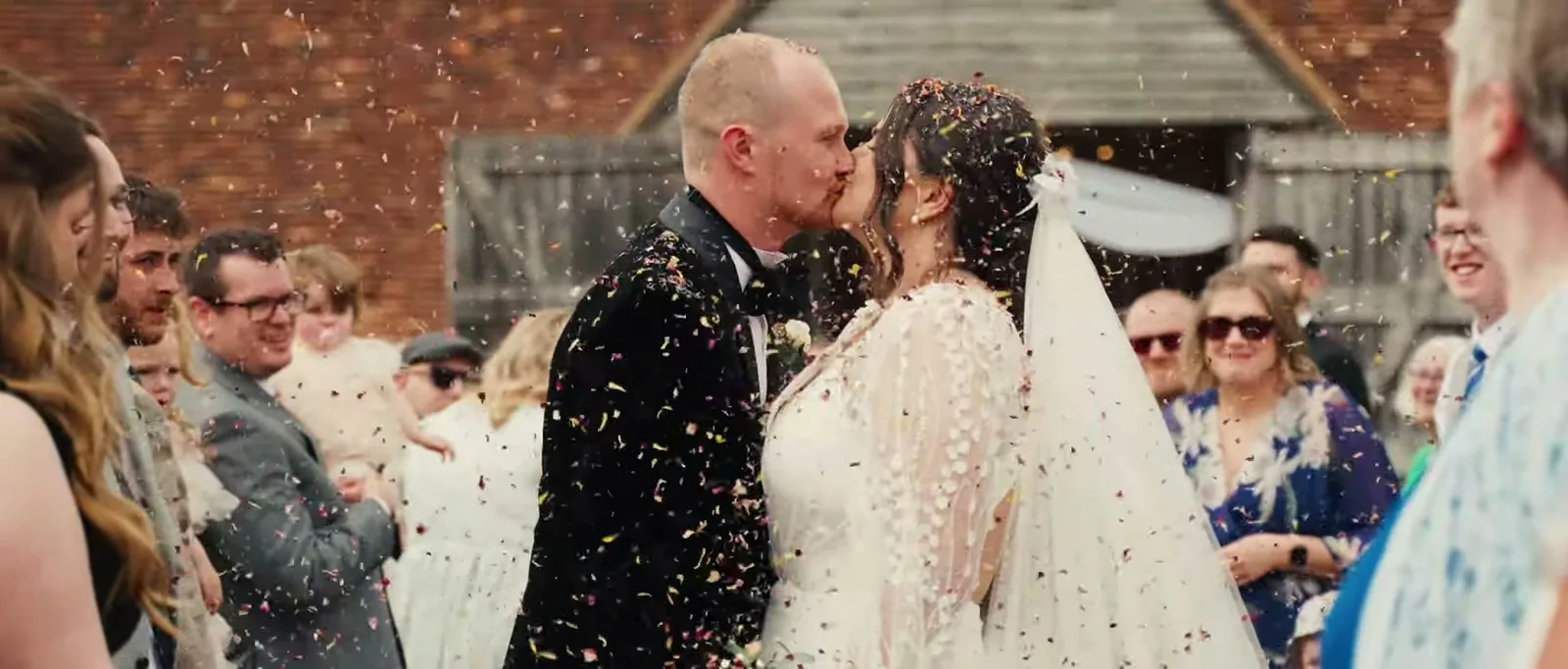 A groom and a bride in a veil kiss in a cloud of petals while smiling guests look on