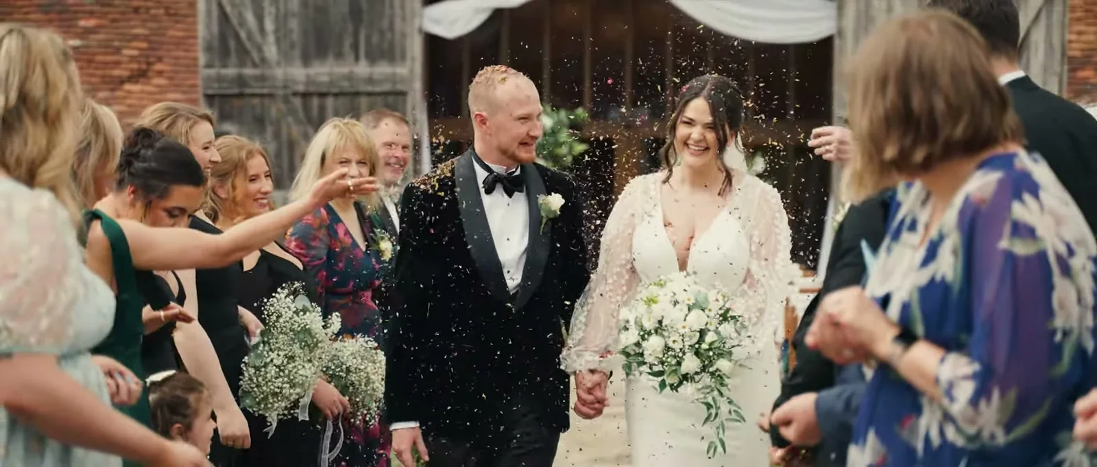 A couple walk hand in hand through a shower of confetti between lines of laughing guests in front of the barn doors
