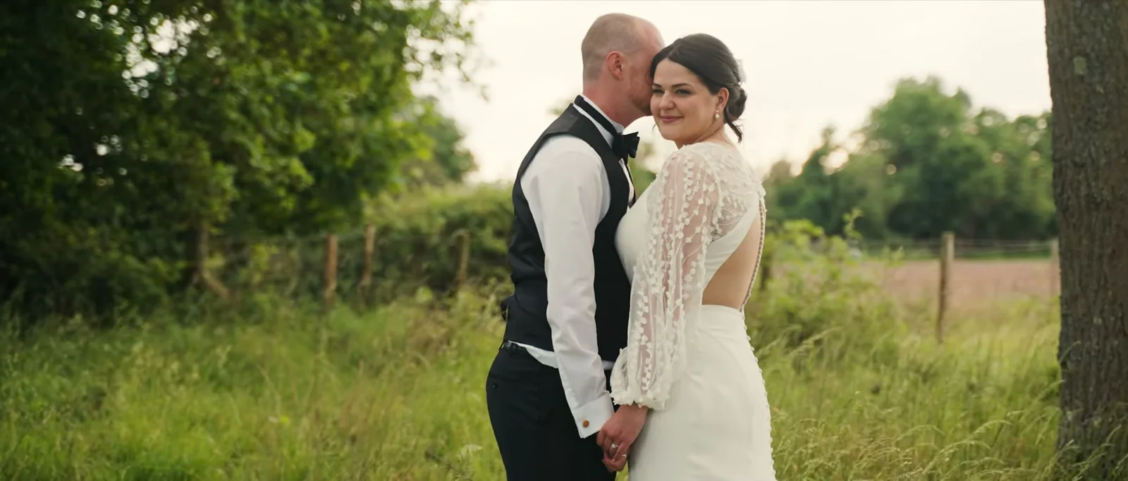A groom kisses the side of his bride's head as she smiles towards the camera, with long grass and a fence behind them