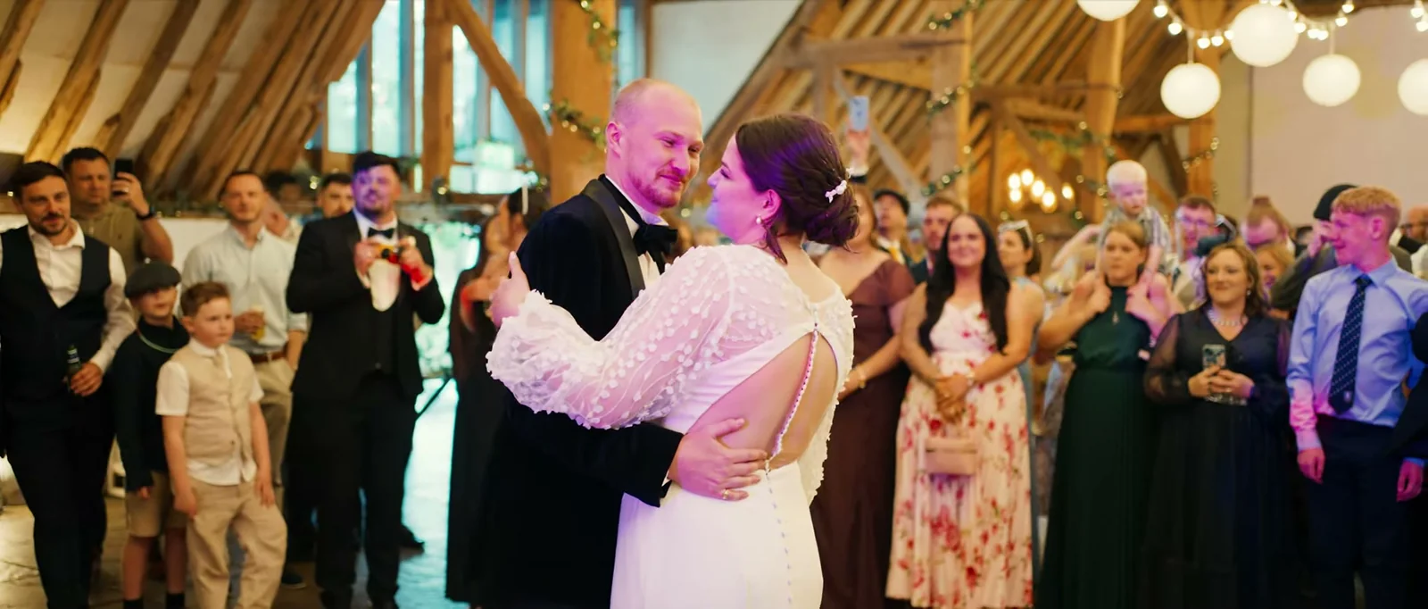 A groom and a bride share their first dance beneath oak beams and white paper lanterns while guests stand around them