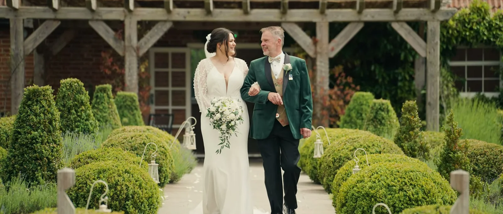 A bride carrying a white bouquet walks on the arm of a man in a green velvet jacket between box hedges and hanging lanterns, both smiling