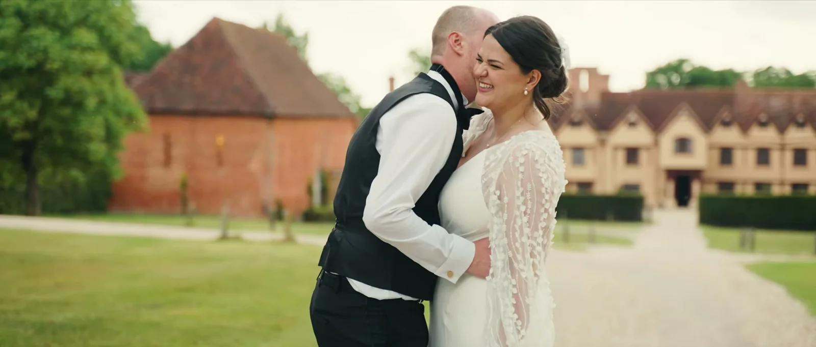 A bride laughs as her groom holds her close on a gravel drive, with a red-brick barn and the manor house blurred behind them