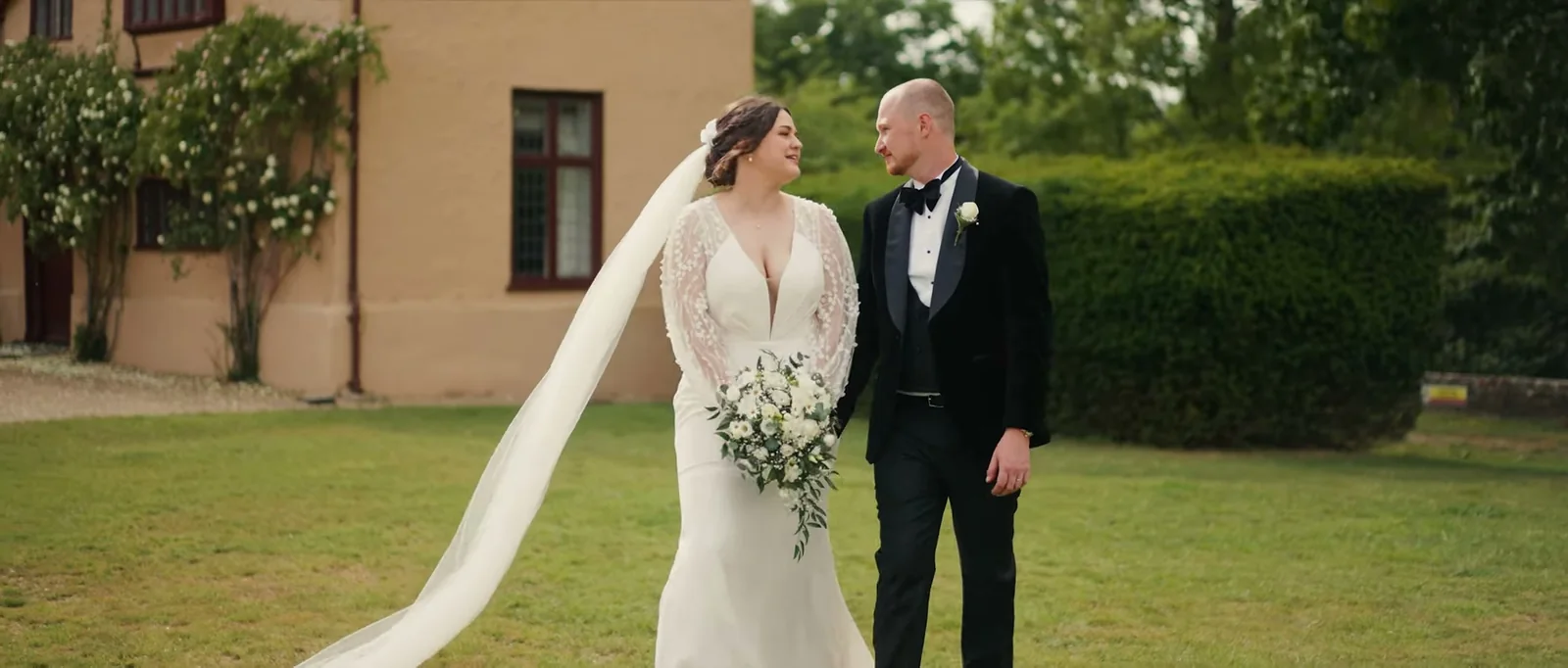 A bride in a long veil and a groom in a black velvet jacket and bow tie walk hand in hand across a lawn beside the pale wall of Ufton Court