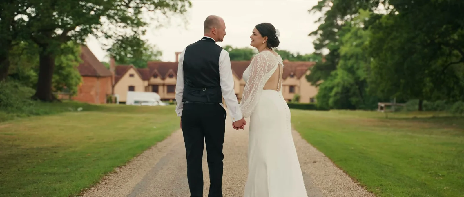 A bride and groom seen from behind hold hands on a gravel drive and look at each other, with the manor house ahead of them