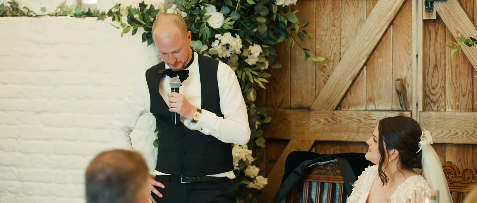 A groom in a waistcoat and bow tie holds a microphone and looks down while his bride watches from the top table in front of wooden barn doors