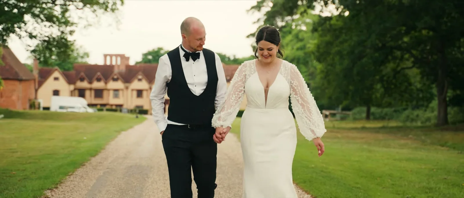 A bride and groom walk hand in hand along a gravel drive towards the manor house, smiling and looking at the ground