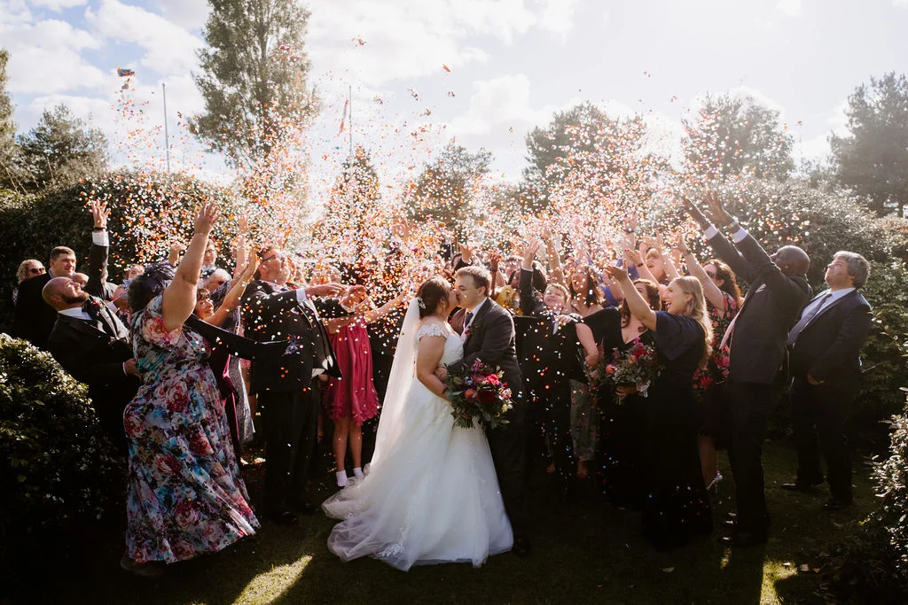 Charlotte and Tom kissing beneath confetti surrounded by guests at Draycote Hotel