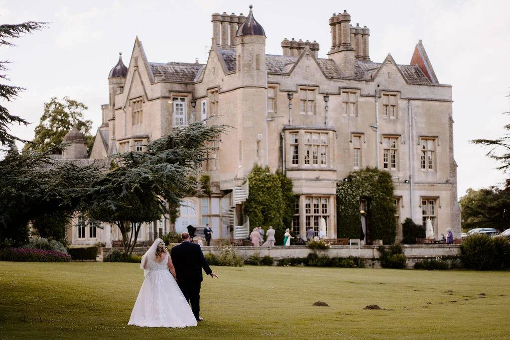 Emma and Jason walking across the lawn with Dumbleton Hall behind them