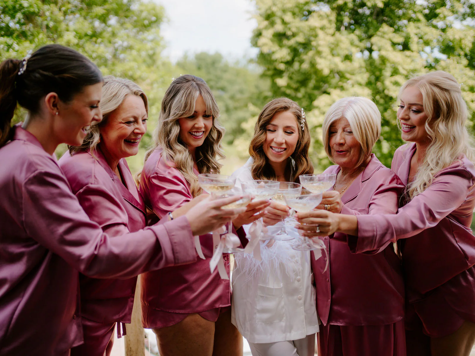 Natalie in a white robe and five women in berry-pink satin pyjamas clink coupe glasses together outdoors beside a wooden structure