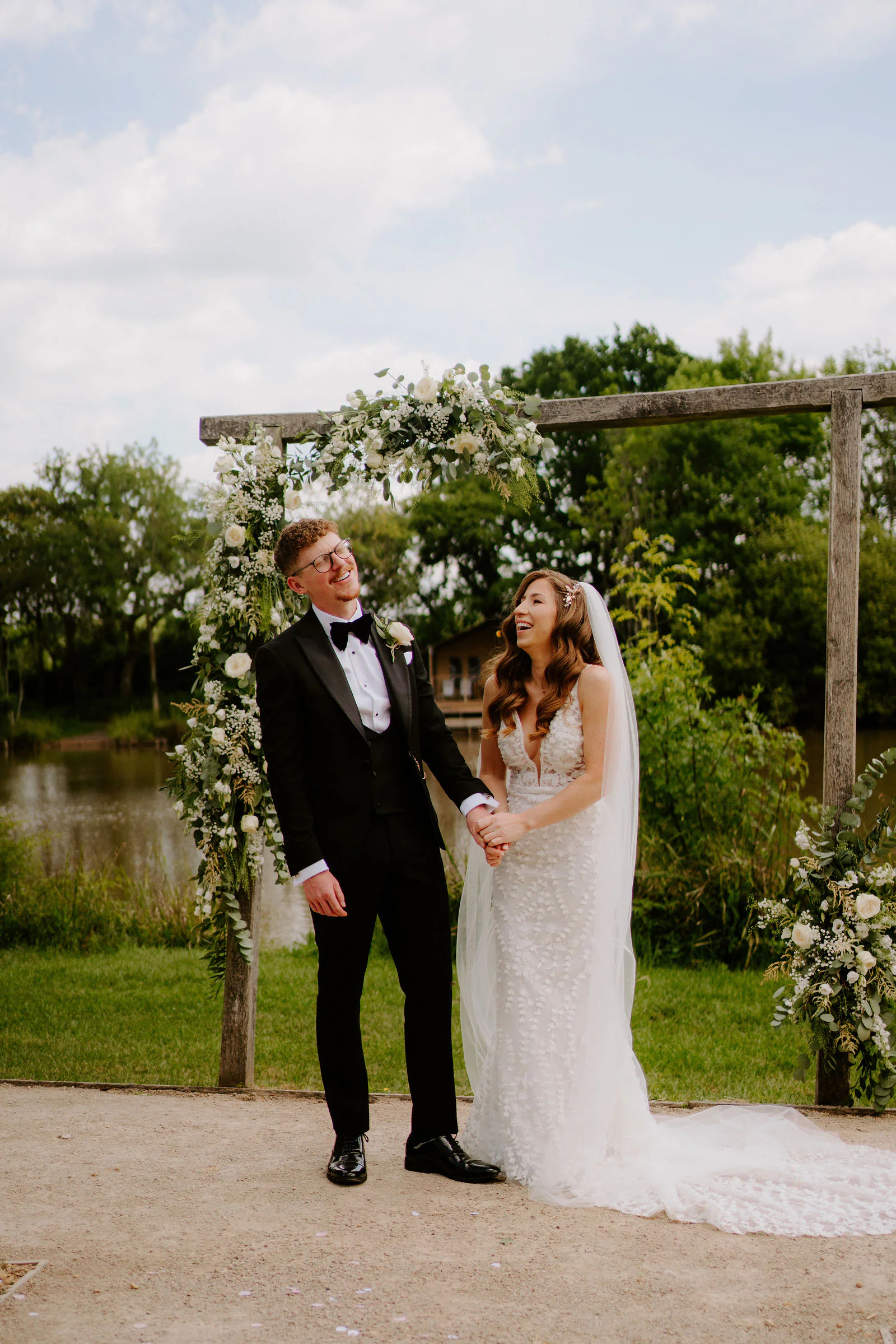 Natalie and Tom stand hand in hand in front of a wooden arch of white flowers beside the lake at Elmbridge Farm
