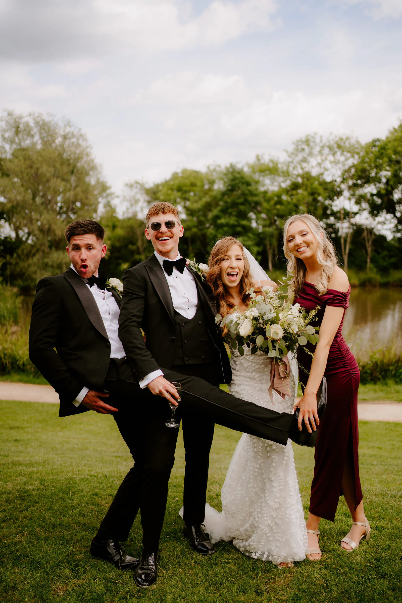 Tom kicks a leg up beside Natalie as a groomsman in sunglasses and a bridesmaid in burgundy laugh with them on the lawn at Elmbridge Farm
