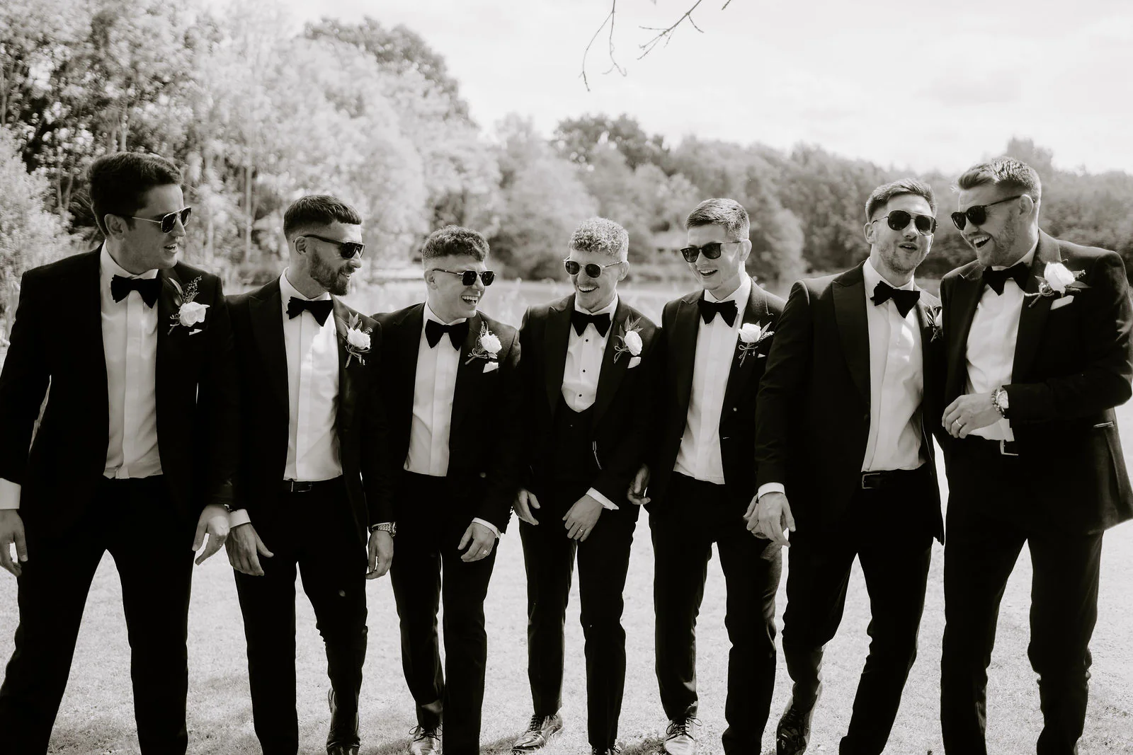 In black and white, seven men in black dinner suits and bow ties, some in sunglasses, walk towards the camera beside the lake at Elmbridge Farm