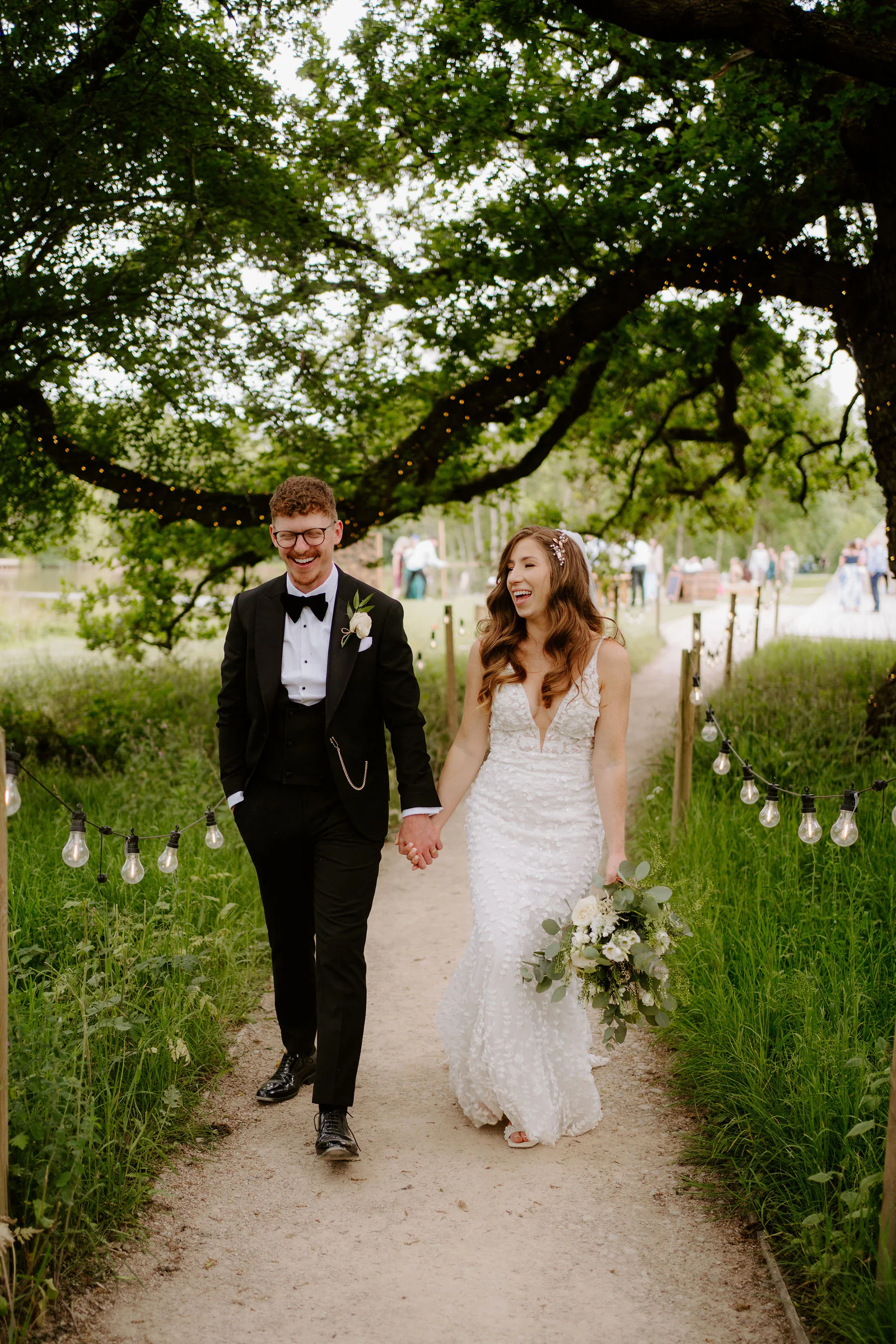 Natalie and Tom walk hand in hand along a path lined with festoon lights beneath a large oak tree at Elmbridge Farm