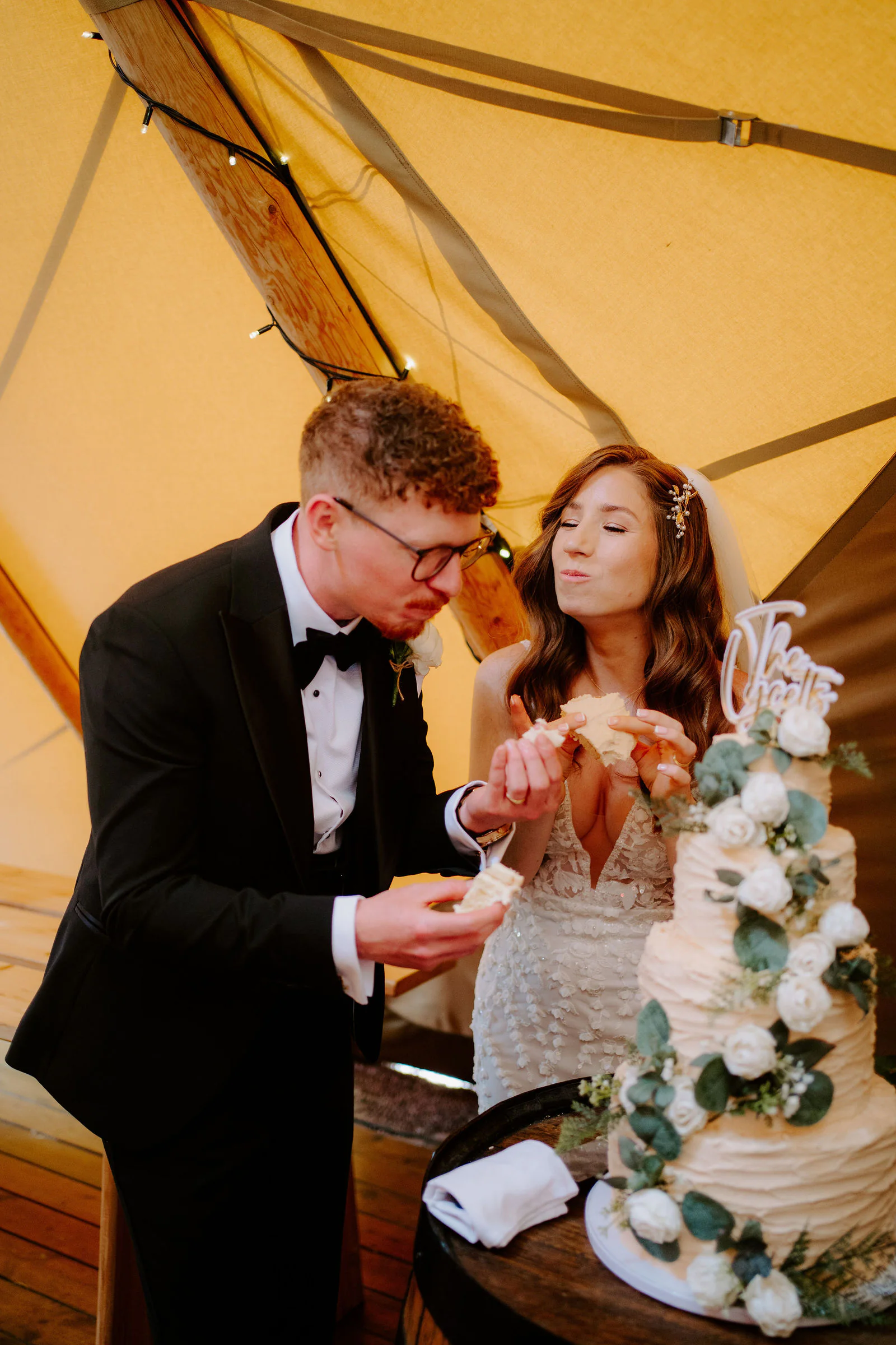Natalie and Tom eat cake beside a four-tier white cake decorated with roses inside a tipi with yellow canvas walls