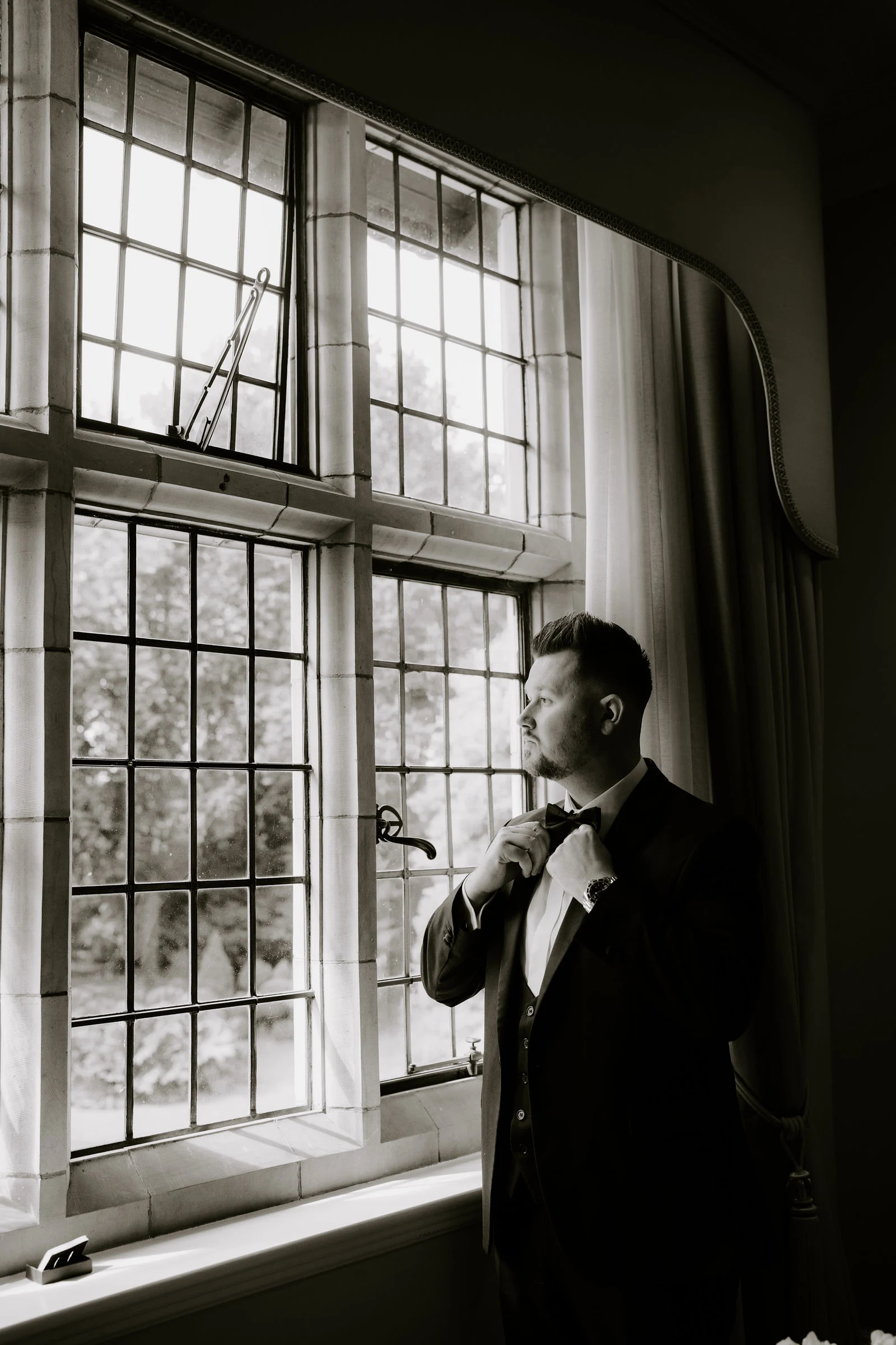 James adjusting his tie beside a large window during preparations at Moxhull Hall