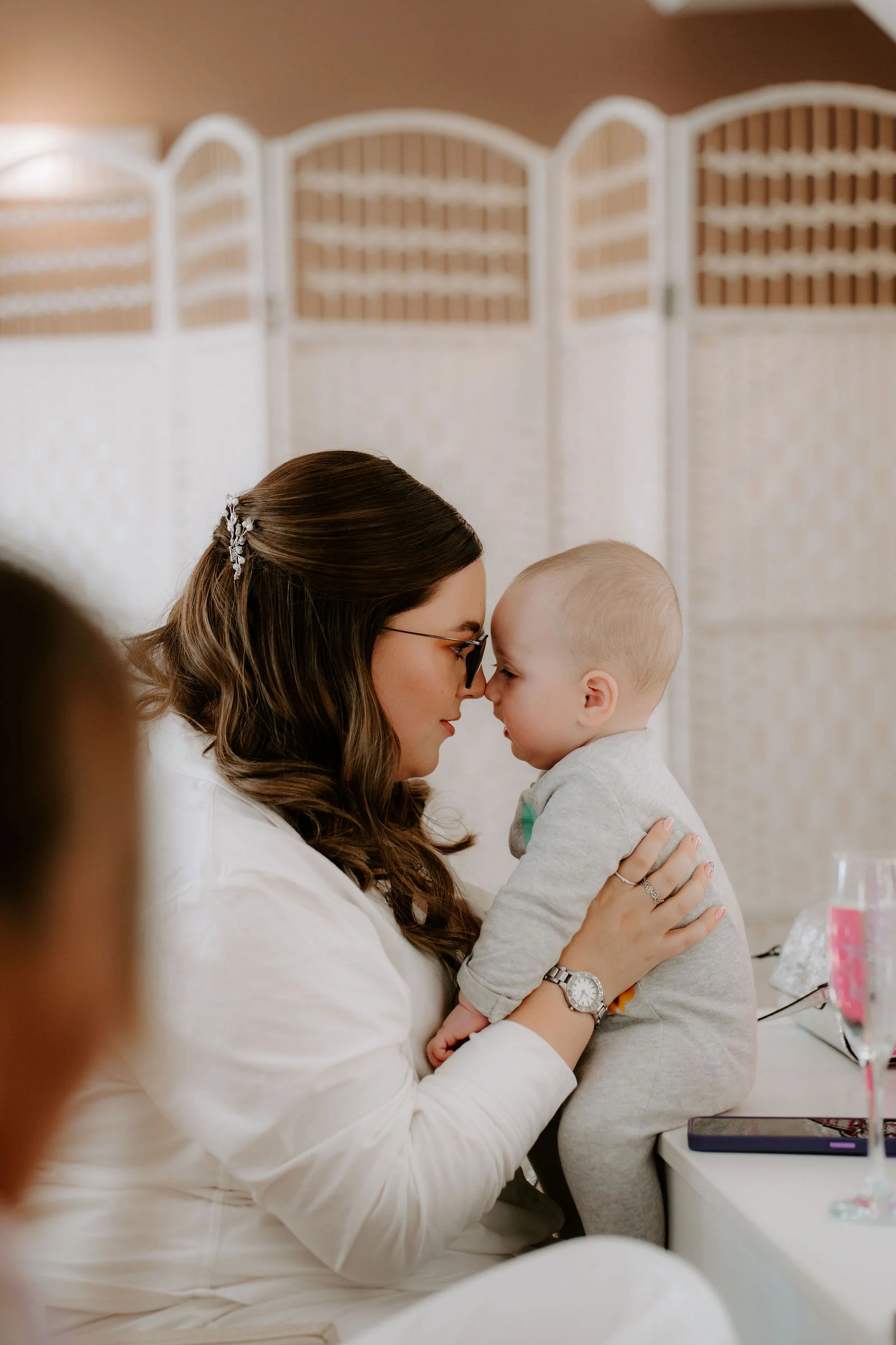 A bride in a white jacket with her hair pinned and glasses presses her nose to a baby's in a bright room with white screens behind her