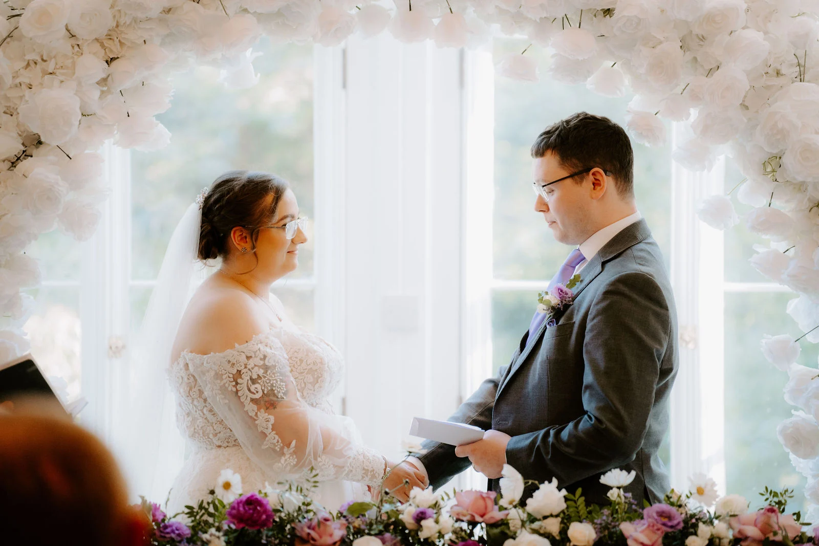 A bride in an off-the-shoulder lace gown and veil and a groom in a grey suit and glasses face each other for their vows in front of tall white windows framed by an arch of white flowers, with pink and purple blooms in front of them
