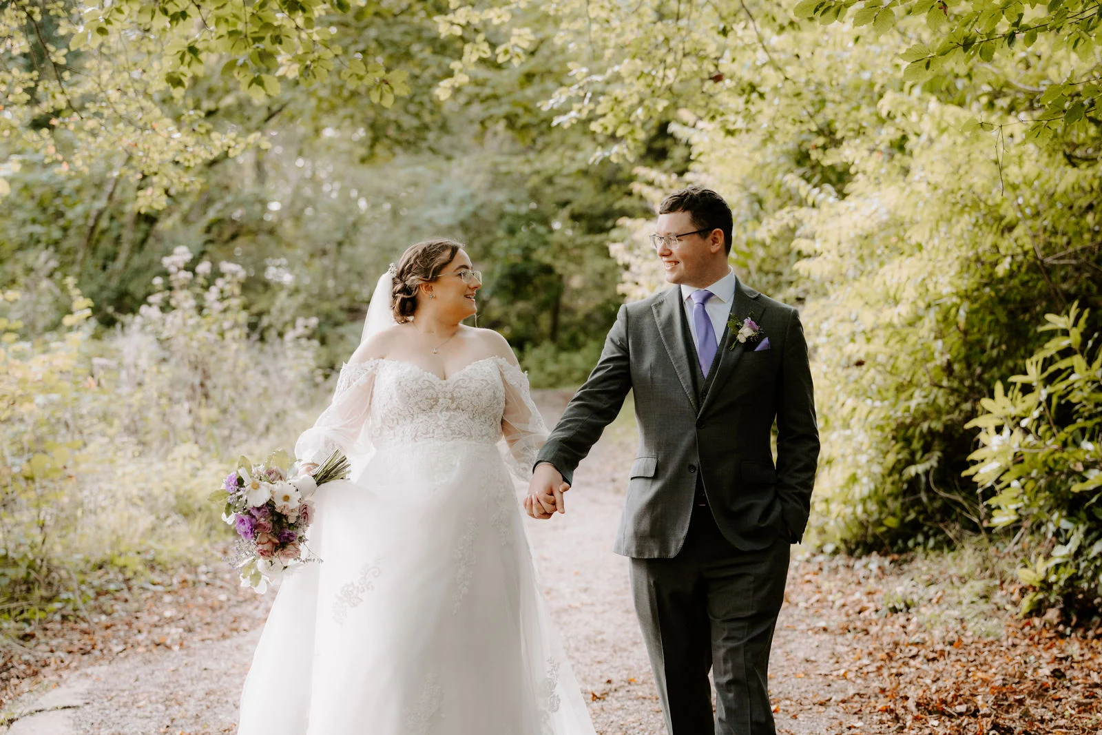 A bride in a white gown with sheer sleeves holding a bouquet of purple and white flowers and a groom in a grey suit with a lilac tie walk hand in hand along a gravel woodland path scattered with autumn leaves
