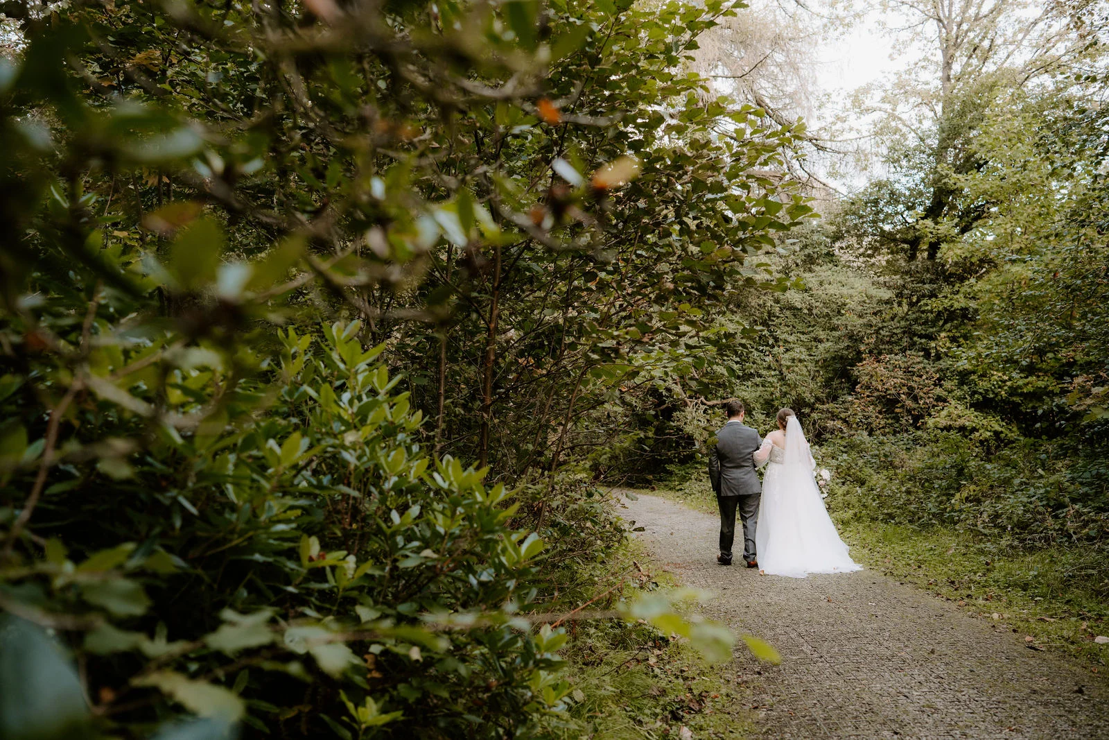 A couple seen from far behind walk along a gravel path through dense woodland, the bride's long white dress trailing behind her