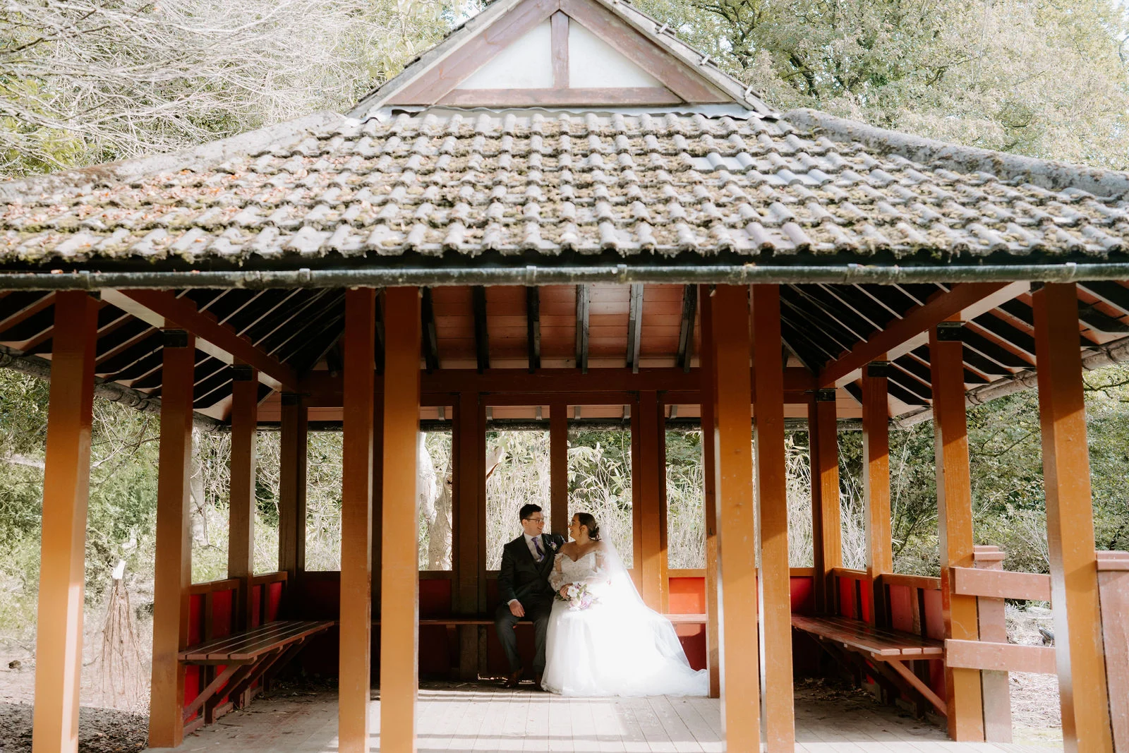 A bride and groom sit together in an open oriental-style timber pavilion with a tiled roof, seen from a distance through its posts