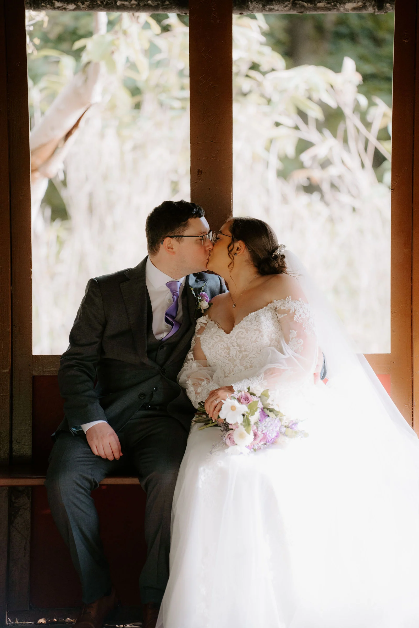 A groom in a grey suit and a bride in a lace gown kiss as they sit together in a window opening of the timber pavilion, her bouquet in her lap