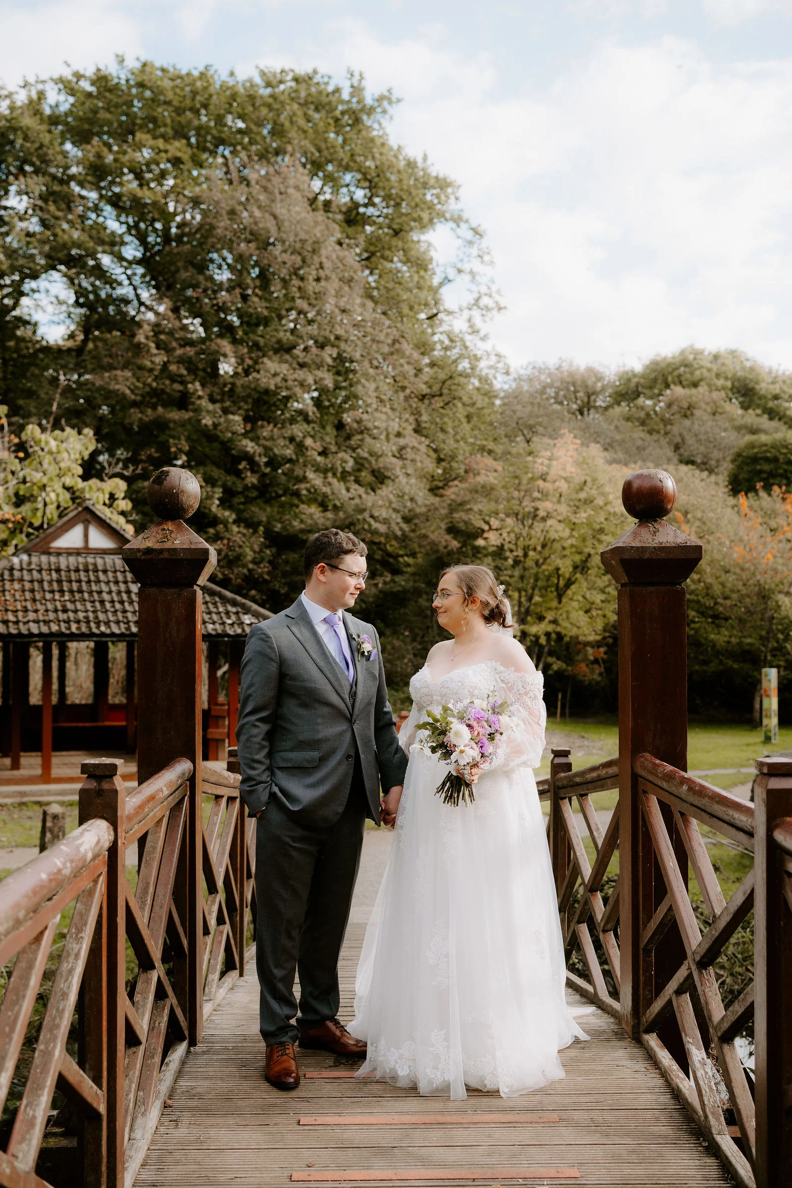 A bride in a white gown and a groom in a grey suit and lilac tie stand hand in hand on a wooden bridge with carved posts, a tile-roofed pavilion and autumn trees behind them