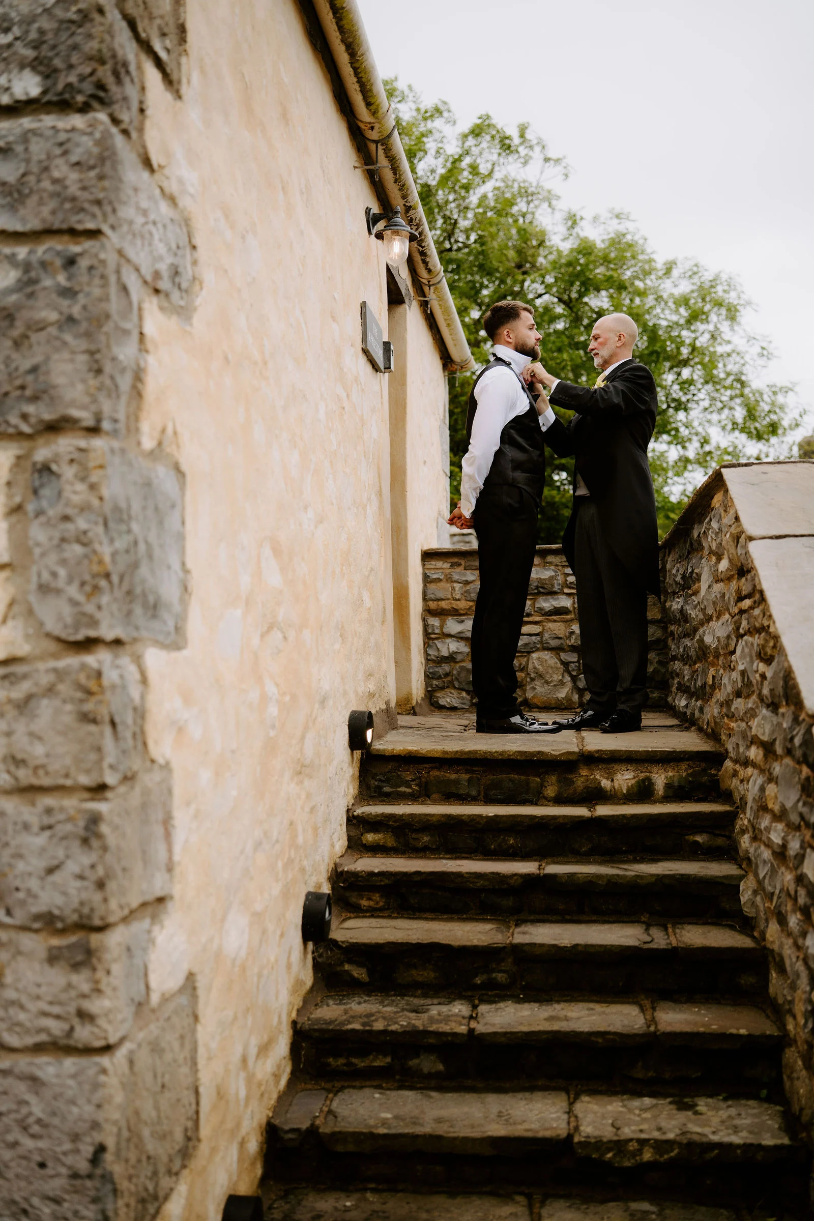 Two men in black waistcoats adjust a bow tie on a stone landing at the top of a flight of old stone steps beside a rough stone wall