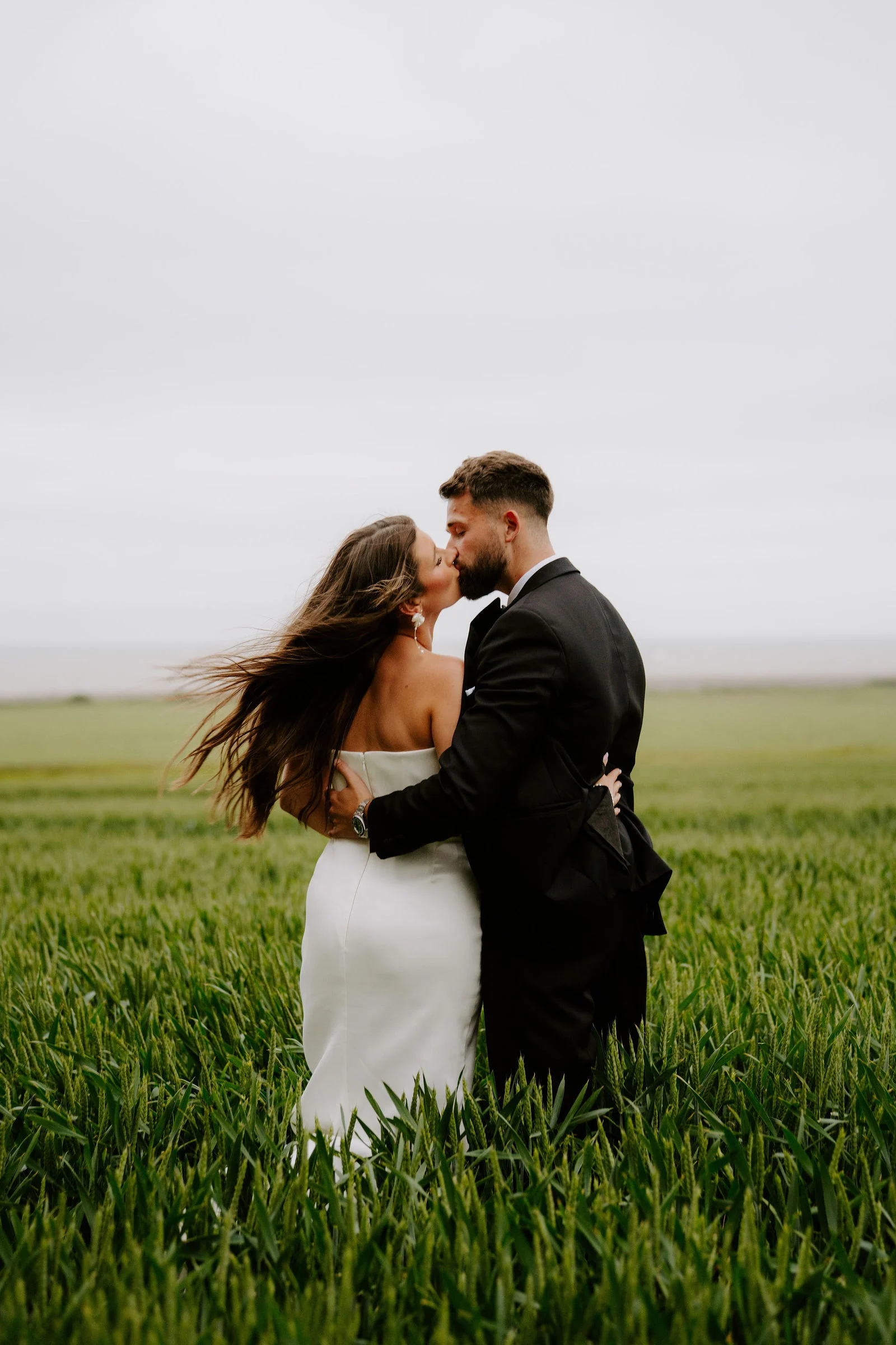A bride with windblown hair and a groom in a dark suit kiss in a field of bright green wheat under a grey sky