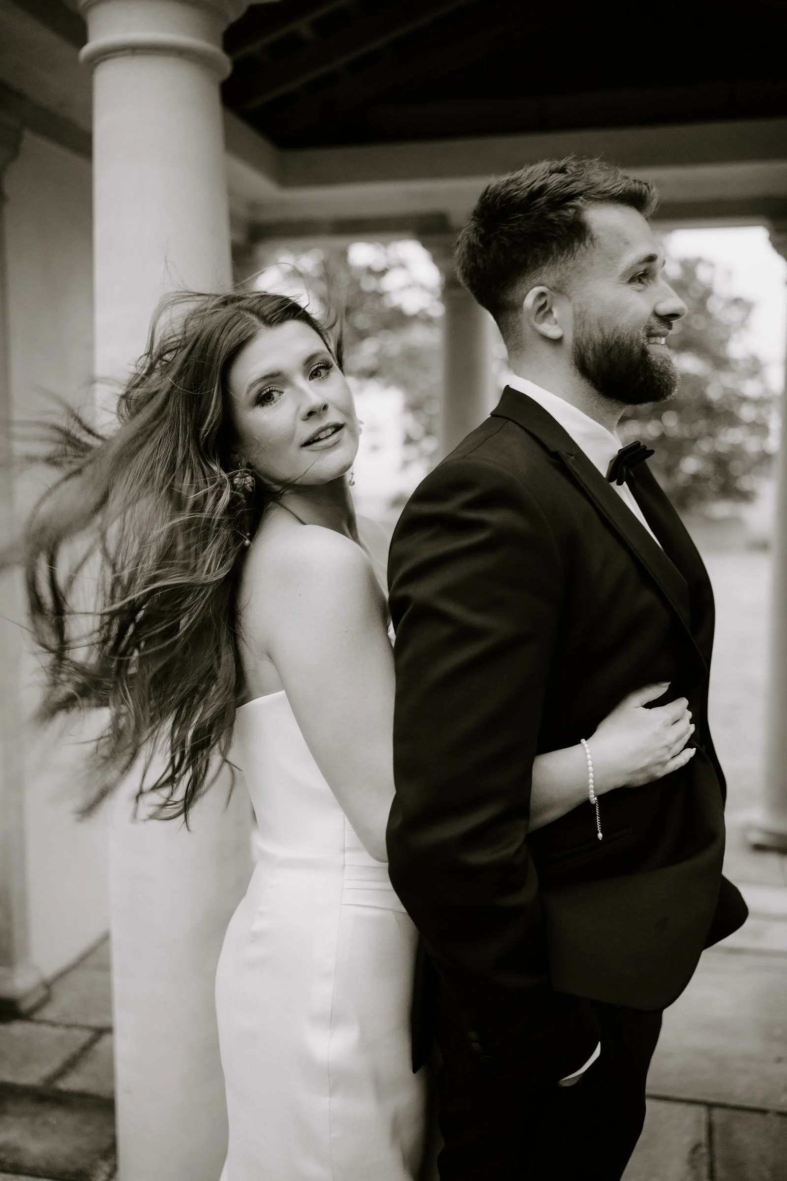 In black and white, a bride with windblown hair glances over her shoulder as her groom in a dinner suit and bow tie holds her from behind between stone pillars