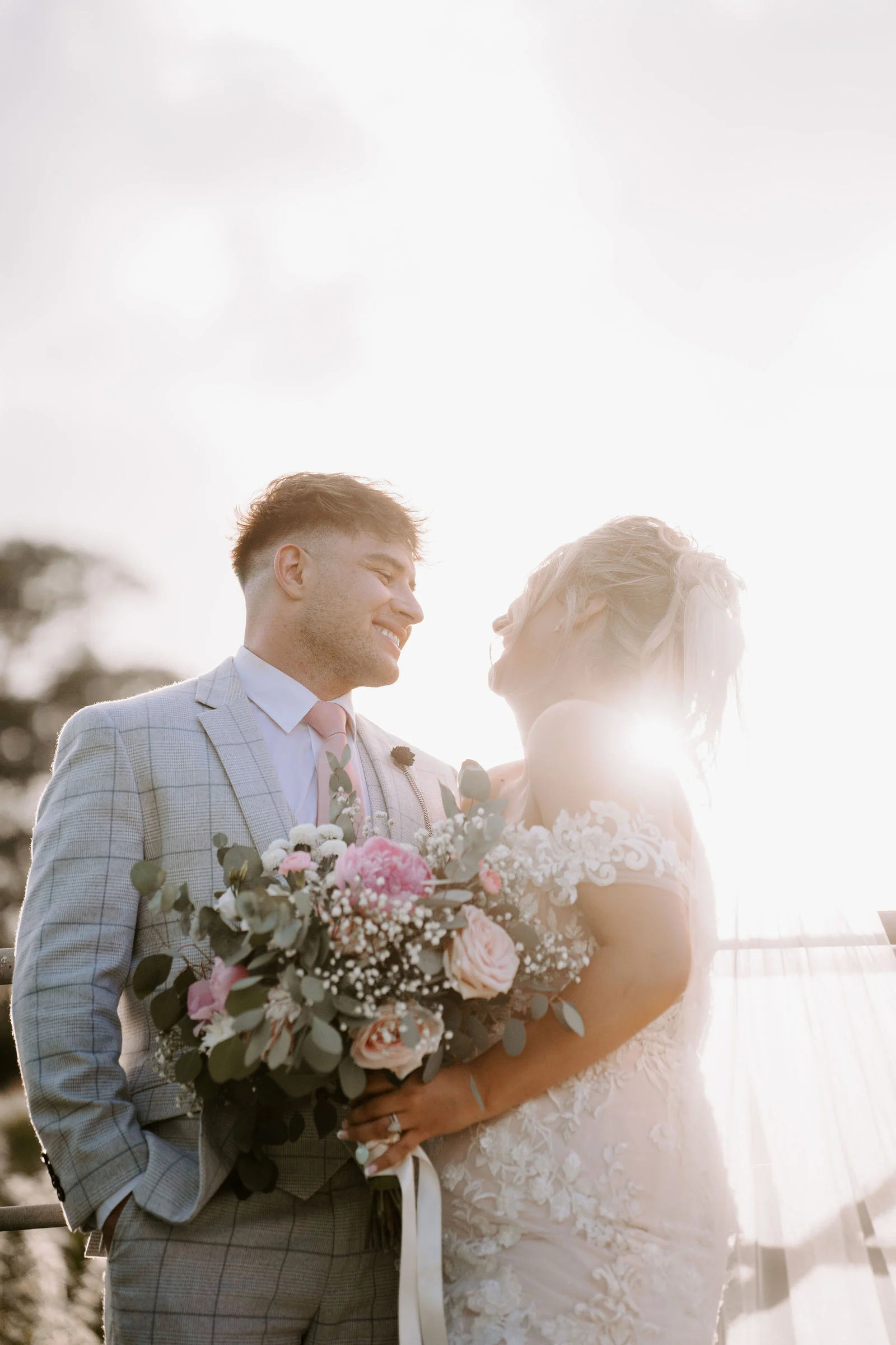A groom in a light check suit holds a pink and white bouquet while his bride in lace smiles at him, both lit by warm backlit sun