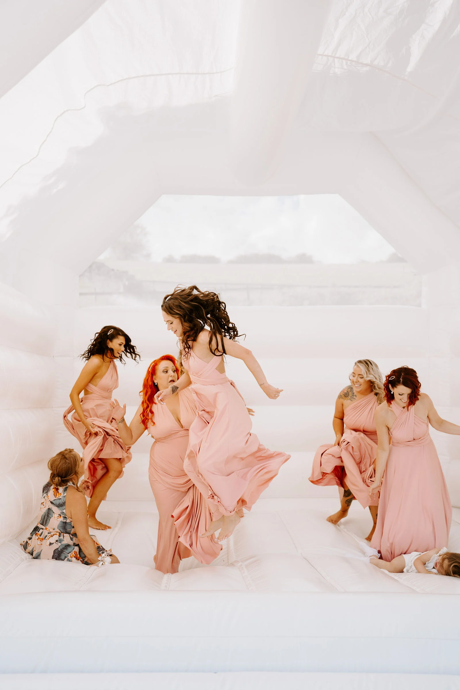 A group of bridesmaids and children in pink dresses jump and dance on a white bouncy castle