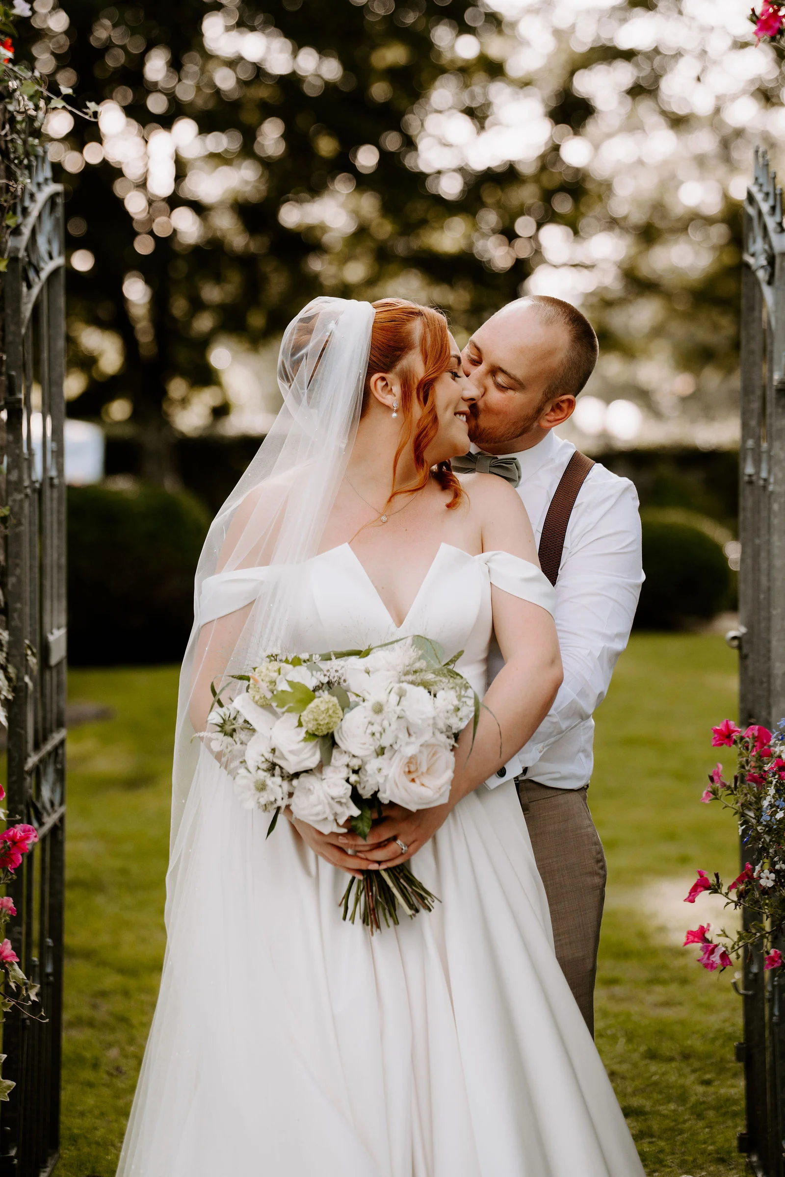 A bride holding a bouquet of white roses is held from behind by her groom as they kiss in an open garden gate edged with pink flowers, with a lawn behind them