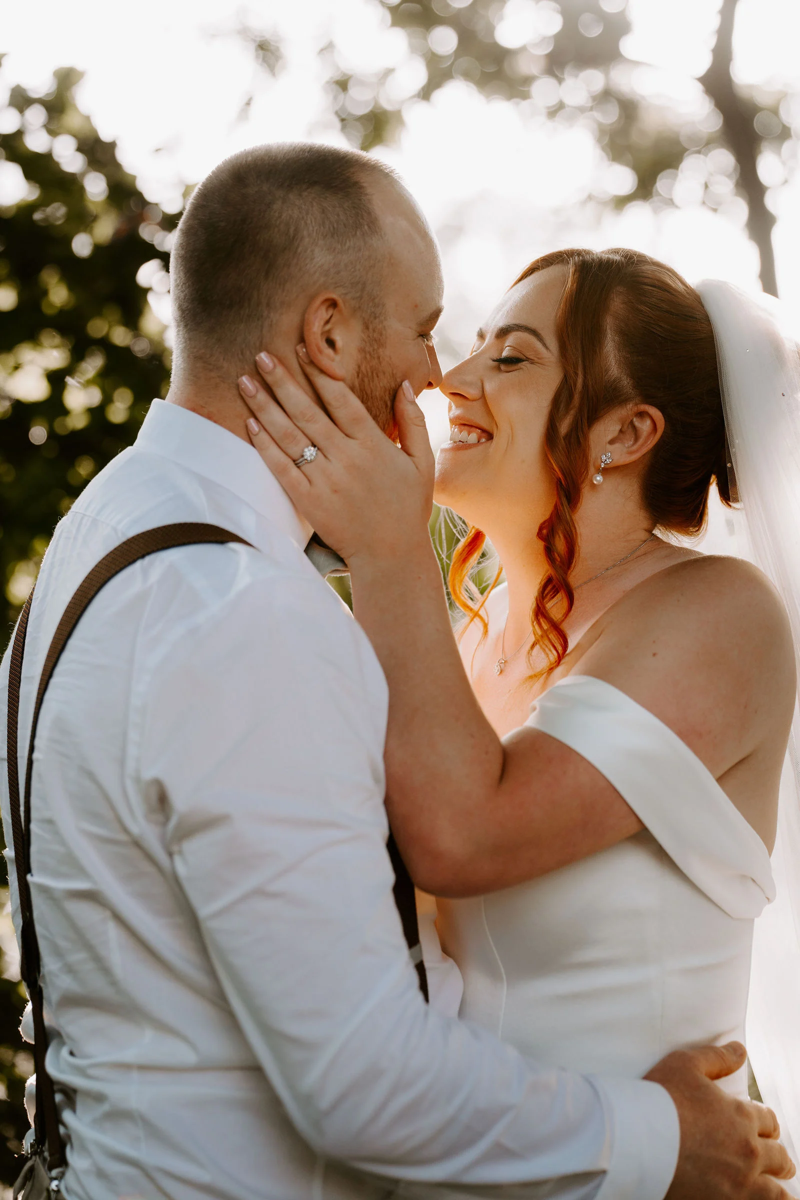 A bride in an off-the-shoulder gown laughs with a hand on her groom's cheek while he holds her close, in warm backlit evening light